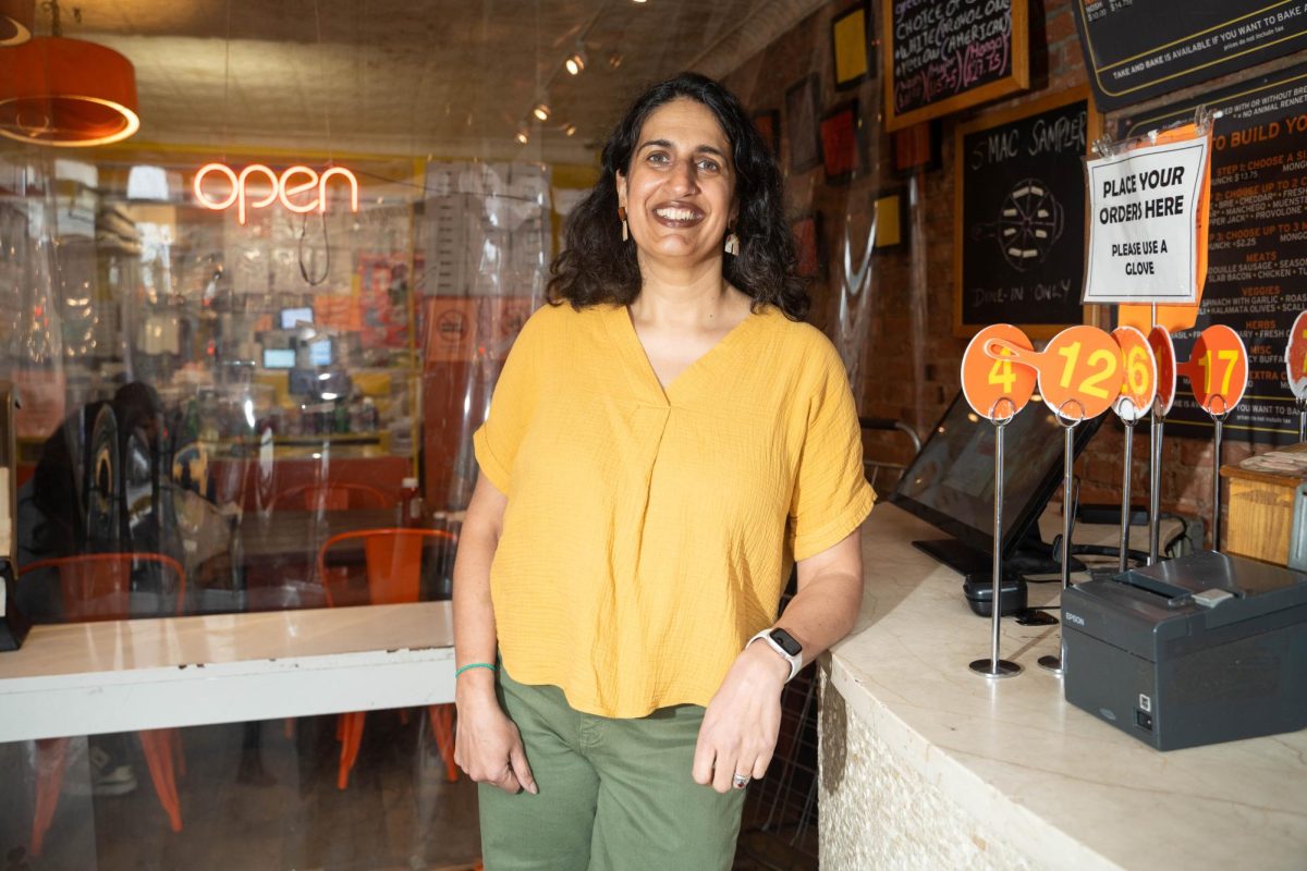 A woman leans on the front counter of a restaurant. She is wearing a yellow shirt and green pants.